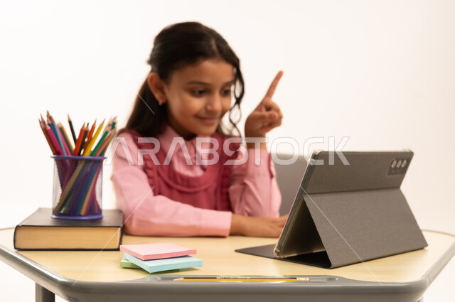 متابعة حصص الاونلاين عن طريق الانترنت, استخدام التكنولوجيا الحديثة، مفهوم التعلم عن بعد، بورتريه لطالبة عربية خليجية سعودية ترتدي اليونيفووم المدرسي تجلس على طاولة الدراسة   تعمل على جهاز التابلت اللوحي بتعابير السرور، خلفية بيضاء