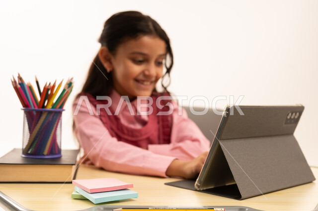مفهوم التعلم عن بعد, متابعة حصص الاونلاين عن طريق الانترنت, استخدام التكنولوجيا الحديثة، بورتريه لطالبة عربية خليجية سعودية ترتدي اليونيفووم المدرسي تجلس على طاولة الدراسة   تعمل على جهاز التابلت اللوحي بتعابير السرور، خلفية بيضاء