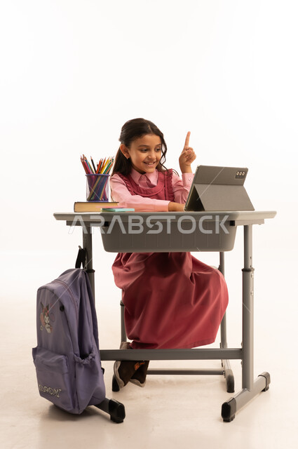 متابعة حصص الاونلاين عن طريق الانترنت, استخدام التكنولوجيا الحديثة، مفهوم التعلم عن بعد، بورتريه لطالبة عربية خليجية سعودية ترتدي اليونيفووم المدرسي تجلس على طاولة الدراسة   تعمل على جهاز التابلت اللوحي بتعابير السرور، خلفية بيضاء