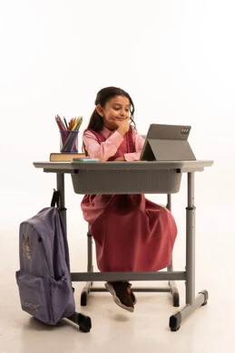 مفهوم التعلم عن بعد, متابعة حصص الاونلاين عن طريق الانترنت, استخدام التكنولوجيا الحديثة، بورتريه لطالبة عربية خليجية سعودية ترتدي اليونيفووم المدرسي تجلس على طاولة الدراسة   تعمل على جهاز التابلت اللوحي بتعابير السرور، خلفية بيضاء