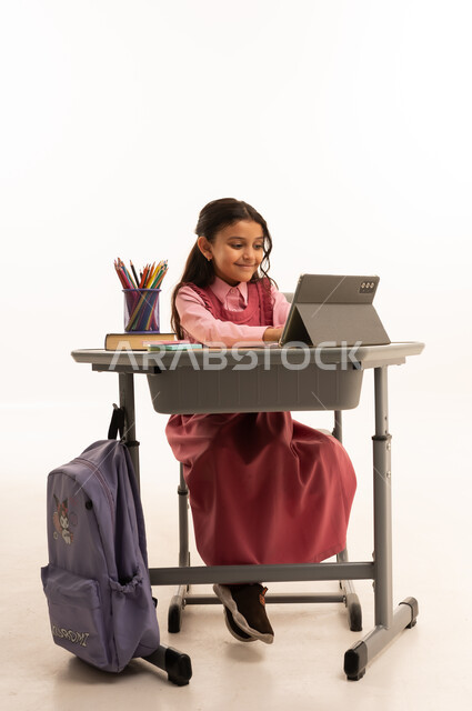 مفهوم التعلم عن بعد, متابعة حصص الاونلاين عن طريق الانترنت, استخدام التكنولوجيا الحديثة، بورتريه لطالبة عربية خليجية سعودية ترتدي اليونيفووم المدرسي تجلس على طاولة الدراسة   تعمل على جهاز التابلت اللوحي بتعابير السرور، خلفية بيضاء