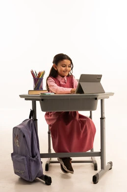 مفهوم التعلم عن بعد, متابعة حصص الاونلاين عن طريق الانترنت, استخدام التكنولوجيا الحديثة، بورتريه لطالبة عربية خليجية سعودية ترتدي اليونيفووم المدرسي تجلس على طاولة الدراسة   تعمل على جهاز التابلت اللوحي بتعابير السرور، خلفية بيضاء