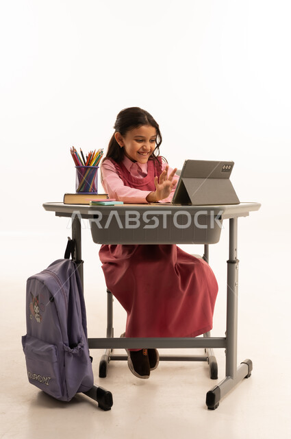 متابعة حصص الاونلاين عن طريق الانترنت, استخدام التكنولوجيا الحديثة، مفهوم التعلم عن بعد، بورتريه لطالبة عربية خليجية سعودية ترتدي اليونيفووم المدرسي تجلس على طاولة الدراسة   تعمل على جهاز التابلت اللوحي بتعابير السرور، خلفية بيضاء