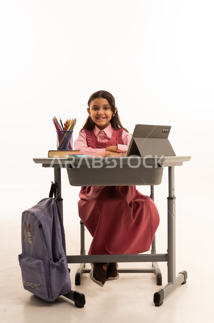 مفهوم التعلم عن بعد, متابعة حصص الاونلاين عن طريق الانترنت, استخدام التكنولوجيا الحديثة، بورتريه لطالبة عربية خليجية سعودية ترتدي اليونيفووم المدرسي تجلس على طاولة الدراسة   تعمل على جهاز التابلت اللوحي بتعابير السرور، خلفية بيضاء