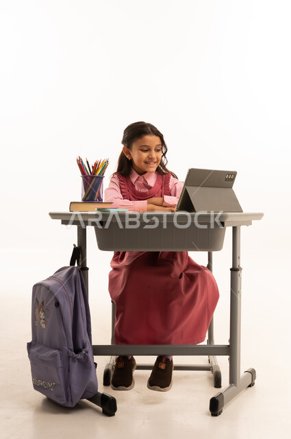 مفهوم التعلم عن بعد, متابعة حصص الاونلاين عن طريق الانترنت, استخدام التكنولوجيا الحديثة، بورتريه لطالبة عربية خليجية سعودية ترتدي اليونيفووم المدرسي تجلس على طاولة الدراسة   تعمل على جهاز التابلت اللوحي بتعابير السرور، خلفية بيضاء