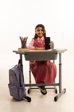 عرض شاشة سوداء فارغة, مفهوم التعلم عن بعد, متابعة حصص الاونلاين عن طريق الانترنت, استخدام التكنولوجيا الحديثة، بورتريه لطالبة عربية خليجية سعودية ترتدي اليونيفووم المدرسي تجلس على طاولة الدراسة   تعمل على الهاتف المحمول بتعابير السرور، خلفية بيضاء