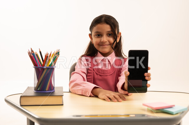 عرض شاشة سوداء فارغة, مفهوم التعلم عن بعد, متابعة حصص الاونلاين عن طريق الانترنت, استخدام التكنولوجيا الحديثة، بورتريه لطالبة عربية خليجية سعودية ترتدي اليونيفووم المدرسي تجلس على طاولة الدراسة   تعمل على الهاتف المحمول بتعابير السرور، خلفية بيضاء