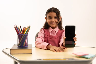 عرض شاشة سوداء فارغة, مفهوم التعلم عن بعد, متابعة حصص الاونلاين عن طريق الانترنت, استخدام التكنولوجيا الحديثة، بورتريه لطالبة عربية خليجية سعودية ترتدي اليونيفووم المدرسي تجلس على طاولة الدراسة   تعمل على الهاتف المحمول بتعابير السرور، خلفية بيضاء