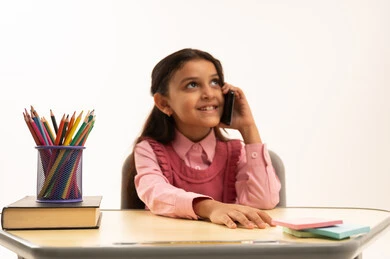 اجراء مكالمة صوتية, مفهوم التعلم عن بعد, متابعة حصص الاونلاين عن طريق الانترنت, استخدام التكنولوجيا الحديثة، بورتريه لطالبة عربية خليجية سعودية ترتدي اليونيفووم المدرسي تجلس على طاولة الدراسة   تعمل على الهاتف المحمول بتعابير السرور، خلفية بيضاء