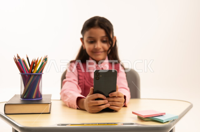 دمج التقنية بالتعليم, مفهوم التعلم عن بعد, متابعة حصص الاونلاين عن طريق الانترنت, استخدام التكنولوجيا الحديثة، بورتريه لطالبة عربية خليجية سعودية ترتدي اليونيفووم المدرسي تجلس على طاولة الدراسة   تعمل على الهاتف المحمول بتعابير السرور، خلفية بيضاء