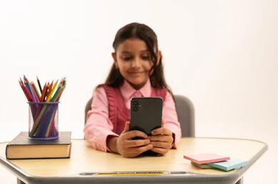 دمج التقنية بالتعليم, مفهوم التعلم عن بعد, متابعة حصص الاونلاين عن طريق الانترنت, استخدام التكنولوجيا الحديثة، بورتريه لطالبة عربية خليجية سعودية ترتدي اليونيفووم المدرسي تجلس على طاولة الدراسة   تعمل على الهاتف المحمول بتعابير السرور، خلفية بيضاء