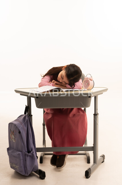 النوم على طاولة الدراسة، ايماءات النعاس والتعب والإرهاق، بورتريه لطالبة عربية خليجية سعودية ترتدي اليونيفورم المدرسي تقوم بإهمال دروسها، سلبيات التعلم عن بعد، هدر وضياع الوقت، خلفية بيضاء