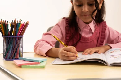 مراجعة الدروس اليومية, مفهوم العودة إلى المدارس، التعليم في المملكة العربية السعودية, الاستعداد للعام الدراسي الجديد, بورتريه مقرب لفتاة عربية خليجية سعودية ترتدي الزي المدرسي تجلس على الطاولة المكتبية تقوم بالكتابة فى الدفتر, خلفية بيضاء