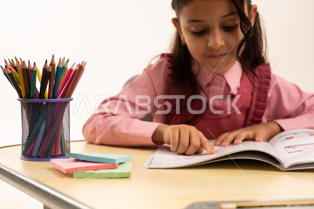 مراجعة الدروس اليومية, مفهوم العودة إلى المدارس، التعليم في المملكة العربية السعودية, الاستعداد للعام الدراسي الجديد, بورتريه مقرب لفتاة عربية خليجية سعودية ترتدي الزي المدرسي تجلس على الطاولة المكتبية تقوم بالكتابة فى الدفتر, خلفية بيضاء
