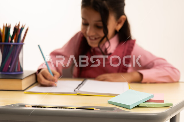 Reviewing daily lessons, back to school concept, education in Saudi Arabia, preparing for the new school year, close-up portrait of a Saudi Arabian Gulf girl wearing school uniform sitting at an office table writing in a notebook, white background