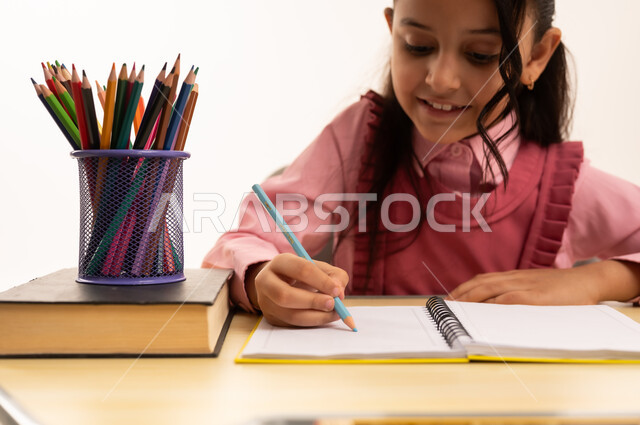 Reviewing daily lessons, back to school concept, education in Saudi Arabia, preparing for the new school year, close-up portrait of a Saudi Arabian Gulf girl wearing school uniform sitting at an office table writing in a notebook, white background