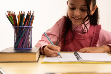 مراجعة الدروس اليومية, مفهوم العودة إلى المدارس، التعليم في المملكة العربية السعودية, الاستعداد للعام الدراسي الجديد, بورتريه مقرب لفتاة عربية خليجية سعودية ترتدي الزي المدرسي تجلس على الطاولة المكتبية تقوم بالكتابة فى الدفتر, خلفية بيضاء