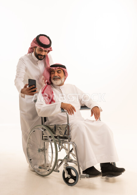 استخدام الهاتف المحمول, توفير اجهزة ومعدات تساعد بالتنقل، بورتريه  لرجل عربي سعودي خليجي يرتدي الشماغ والثوب التقليدي يقوم بمساعدة ابيه من ذوي الاحتياجات الخاصه يجلس على كرسي متحرك، شعور المعاقين بالإنتماء والتقدير والإحترام في السعوديه، خلفيه بيضاء