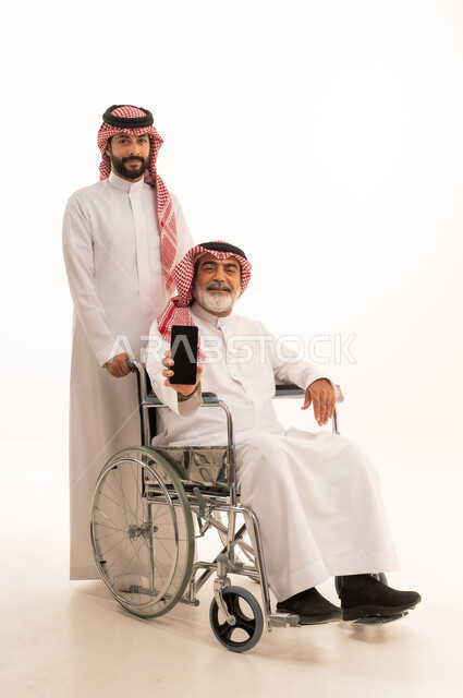عرض شاشة هاتف محمول سوداء فارغة, توفير اجهزة ومعدات تساعد بالتنقل، بورتريه  لرجل عربي سعودي خليجي يرتدي الشماغ والثوب التقليدي يقوم بمساعدة ابيه من ذوي الاحتياجات الخاصه يجلس على كرسي متحرك، شعور المعاقين بالإنتماء والتقدير والإحترام في السعوديه، خلفيه بيضاء