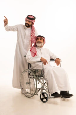  العنايه بأصحاب الهمم وتأمين احتياجاتهم، توفير اجهزة ومعدات تساعد بالتنقل، بورتريه  لرجل عربي سعودي خليجي يرتدي الشماغ والثوب التقليدي يقوم بمساعدة ابيه من ذوي الاحتياجات الخاصه يجلس على كرسي متحرك، شعور المعاقين بالإنتماء والتقدير والإحترام في السعوديه، خلفيه بيضاء