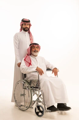 توفير اجهزة ومعدات تساعد بالتنقل، العنايه بأصحاب الهمم وتأمين احتياجاتهم، بورتريه  لرجل عربي سعودي خليجي يرتدي الشماغ والثوب التقليدي يقوم بمساعدة ابيه من ذوي الاحتياجات الخاصه يجلس على كرسي متحرك، شعور المعاقين بالإنتماء والتقدير والإحترام في السعوديه، خلفيه بيضاء
