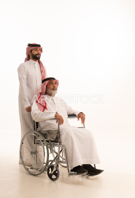  العنايه بأصحاب الهمم وتأمين احتياجاتهم، توفير اجهزة ومعدات تساعد بالتنقل، بورتريه  لرجل عربي سعودي خليجي يرتدي الشماغ والثوب التقليدي يقوم بمساعدة ابيه من ذوي الاحتياجات الخاصه يجلس على كرسي متحرك، شعور المعاقين بالإنتماء والتقدير والإحترام في السعوديه، خلفيه بيضاء