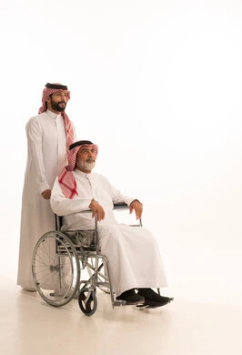 توفير اجهزة ومعدات تساعد بالتنقل، العنايه بأصحاب الهمم وتأمين احتياجاتهم، بورتريه  لرجل عربي سعودي خليجي يرتدي الشماغ والثوب التقليدي يقوم بمساعدة ابيه من ذوي الاحتياجات الخاصه يجلس على كرسي متحرك، شعور المعاقين بالإنتماء والتقدير والإحترام في السعوديه، خلفيه بيضاء