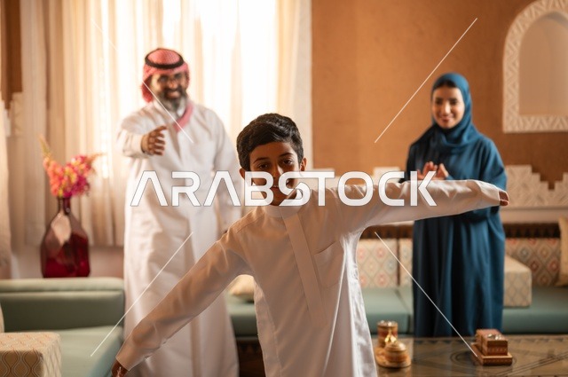 طفل يمثل حركة الطير جو عائلي دافئ مفعم بالحيوية، إظهار مشاعر المودة والمحبة العائلية, عائلة عربية خليجية سعودية تقف داخل غرفة تراثية بديكورات جدران عصرية، تفاصيل المكان المزخرف مع لمسات ديكور تراثية عربية ومزهرية تحتوي على ورود ملونة، التسلية في وقت الفراغ