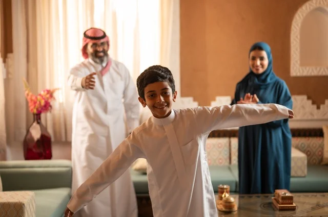 طفل يمثل حركة الطير جو عائلي دافئ مفعم بالحيوية، إظهار مشاعر المودة والمحبة العائلية, عائلة عربية خليجية سعودية تقف داخل غرفة تراثية بديكورات جدران عصرية، تفاصيل المكان المزخرف مع لمسات ديكور تراثية عربية ومزهرية تحتوي على ورود ملونة، التسلية في وقت الفراغ