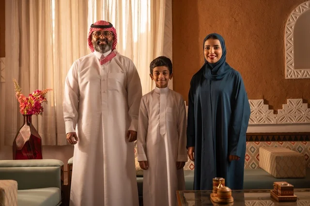 Details of the decorated place with decorative touches and a vase containing colorful flowers, support for the children from the parents, showing feelings of affection and family love, an Arab Gulf Saudi family standing inside a heritage room with modern wall decorations, details of the decorated place with touches of Arab heritage decor.