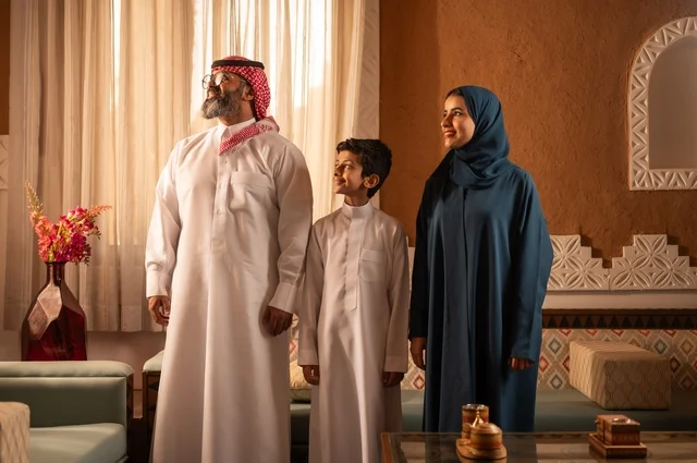 التأمل في مشهد خارجي، إظهار مشاعر المودة والمحبة العائلية, عائلة عربية خليجية سعودية تقف داخل غرفة تراثية بديكورات جدران مزخرفة، تفاصيل المكان المزخرف مع لمسات ديكور تراثية عربية ومزهرية تحتوي على ورود ملونة، التسلية في وقت الفراغ