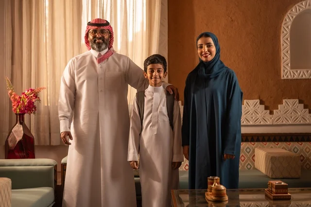 التقاط صورة عائلية بعد العودة من المدرسة، دعم الأولاد من قبل الأبوين، إظهار مشاعر المودة والمحبة العائلية, عائلة عربية خليجية سعودية تقف داخل غرفة تراثية بديكورات جدران مزخرفة,  في الخلفية تفاصيل المكان المزخرف مع لمسات ديكور تراثية عربية