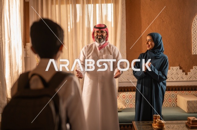 Support for children from parents, at the time of returning from school, showing feelings of affection and family love. A Saudi Gulf Arab family stands inside a heritage room with decorated walls. In the background, there are details of the decorated place with touches of Arab heritage decor.