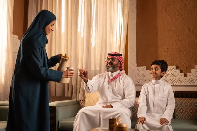 تعليم العادات والتقاليد كرم الضيافة، القهوة العربية تراث أصيل، إظهار مشاعر المودة والمحبة العائلية, صورة لعائلة عربية خليجية سعودية تجلس داخل غرفة تراثية بديكورات جدران مزخرفة,  في الخلفية تفاصيل المكان المزخرف مع لمسات ديكور تراثية عربية