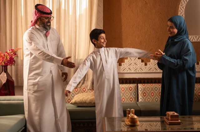 Childish movements bring joy to the parents, expressing feelings of affection and family love. A Saudi Gulf Arab family stands inside a heritage room with modern wall decorations, the details of the decorated place with touches of Arab heritage decor and a vase containing colorful flowers, entertainment in free time.