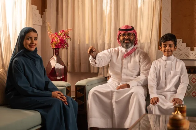 النظر الى الكاميرا بايماءات السعادة ، إظهار مشاعر المودة والمحبة العائلية,صورة لعائلة عربية خليجية سعودية تجلس داخل غرفة تراثية بديكورات جدران مزخرفة,  في الخلفية تفاصيل المكان المزخرف مع لمسات ديكور تراثية عربية، قضاء  وقت العطلة معا