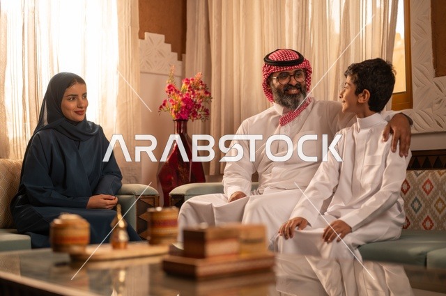 تبادل الأحاديث والمواقف المضجكة مع العائلة ، إظهار مشاعر المودة والمحبة العائلية,صورة لعائلة عربية خليجية سعودية تجلس داخل غرفة تراثية بديكورات جدران مزخرفة,  في الخلفية تفاصيل المكان المزخرف مع لمسات ديكور تراثية عربية، قضاء  وقت العطلة معا