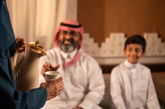 Arabic coffee is an authentic heritage of hospitality, teaching customs and traditions, and expressing feelings of affection and family love. A close-up image of a Saudi Gulf Arab family sitting inside a traditional room with decorated walls. In the background, details of the ornate place with touches of traditional Arab decor.