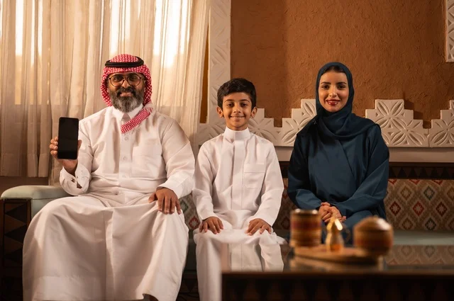 يعرض الأب خلفية شاشة سوداء على الهاتف المحمول، الانسجام بين أفراد الأسرة ،إظهار مشاعر المودة والمحبة العائلية, صورة مقربة لعائلة عربية خليجية سعودية تجلس داخل غرفة تراثية بديكورات جدران مزخرفة,  في الخلفية تفاصيل المكان المزخرف مع لمسات ديكور تراثية عربية
