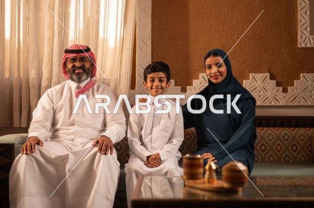 النظر الى الكاميرا بايماءات السعادة ، إظهار مشاعر المودة والمحبة العائلية,صورة لعائلة عربية خليجية سعودية تجلس داخل غرفة تراثية بديكورات جدران مزخرفة,  في الخلفية تفاصيل المكان المزخرف مع لمسات ديكور تراثية عربية، قضاء  وقت العطلة معا