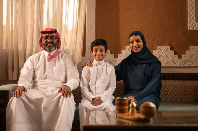 النظر الى الكاميرا بايماءات السعادة ، إظهار مشاعر المودة والمحبة العائلية,صورة لعائلة عربية خليجية سعودية تجلس داخل غرفة تراثية بديكورات جدران مزخرفة,  في الخلفية تفاصيل المكان المزخرف مع لمسات ديكور تراثية عربية، قضاء  وقت العطلة معا