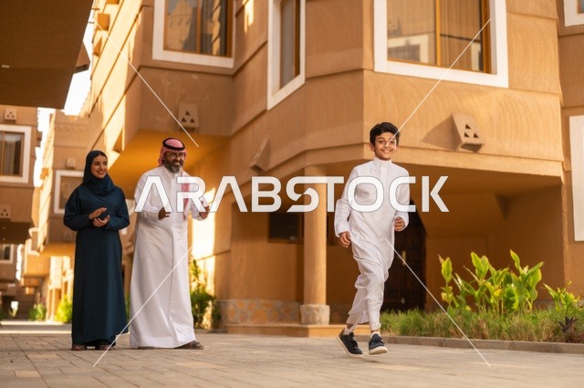 Family recreational activities and events, a family atmosphere filled with love and affection, encouragement and support from parents, a Saudi Gulf Arab family standing at the entrance of a modern home, the father wearing a white thobe with a ghutrah and agal, the child wearing a traditional white thobe, and the mother wearing an abaya and hijab.