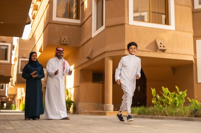 أنشطة وفعاليات ترفيهية عائلة، أجواء أسرية مليئة بالحب والحنان، تشجيع ودعم الوالدين،عائلة عربية خليجية سعودية يقفون في مدخل منزل حديث، يرتدي الأب الثوب الأبيض مع الغترة والعقال، يرتدي الطفل ثوبًا أبيض تقليديًا، أم ترتدي عباءة وحجاب