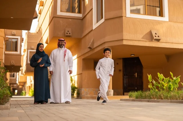 جو العائلة الخليجية والترابط الأسري، أجواء أسرية مليئة بالحب والحنان، تشجيع ودعم الوالدين،عائلة عربية خليجية سعودية يقفون في مدخل منزل حديث، يرتدي الأب الثوب الأبيض مع الغترة والعقال، يرتدي الطفل ثوبًا أبيض تقليديًا، أم ترتدي عباءة وحجاب
