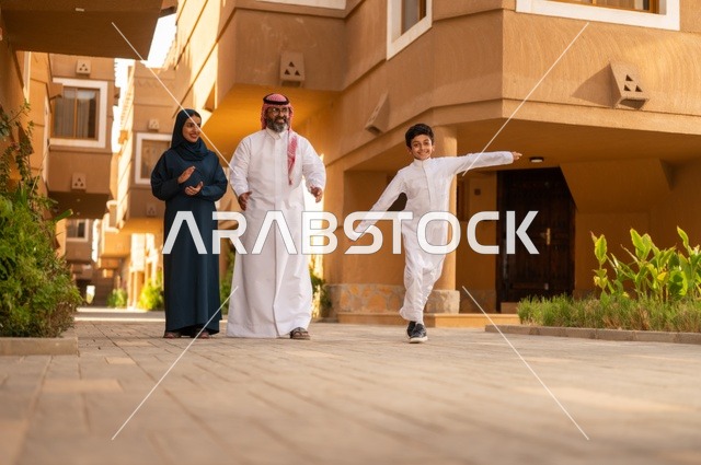 أجواء أسرية مليئة بالحب والحنان، تشجيع ودعم الوالدي،ن جو العائلة الخليجية والترابط الأسري، عائلة عربية خليجية سعودية يقفون في مدخل منزل حديث، يرتدي الأب الثوب الأبيض مع الغترة والعقال، يرتدي الطفل ثوبًا أبيض تقليديًا، أم ترتدي عباءة وحجاب