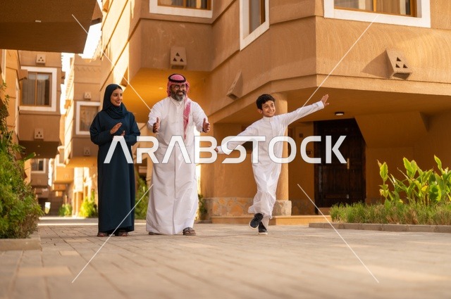 Encouragement and support from parents, a family atmosphere filled with love and tenderness, the environment of the Gulf family and family bonding, an Arab Gulf Saudi family standing at the entrance of a modern home, the father wearing a white thobe with a ghutrah and agal, the child wearing a traditional white thobe, and the mother wearing an abaya and hijab.
