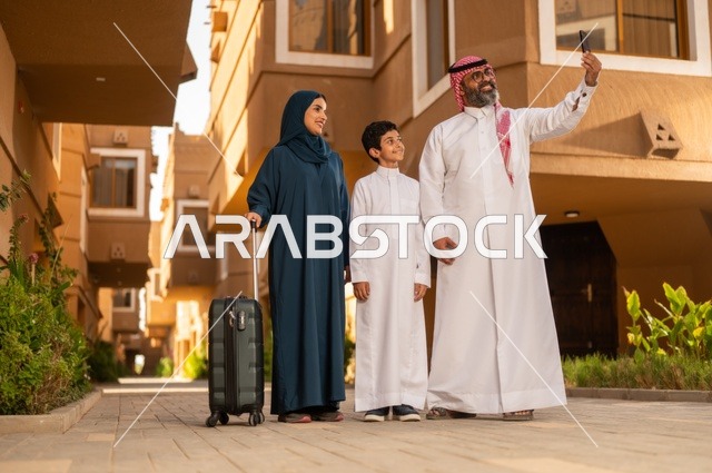 التقاط صورة تذكارية سيلفي للحظات ماقبل السفر، قضاء رحلة صيفية مسلية، جو العائلة الخليجية والترابط الأسري، عائلة عربية خليجية سعودية يقفون في مدخل منزل حديث، يرتدي الأب الثوب الأبيض مع الغترة والعقال، يرتدي الطفل ثوبًا أبيض تقليديًا، تمسك الأم  بيدها حقيبة سفر