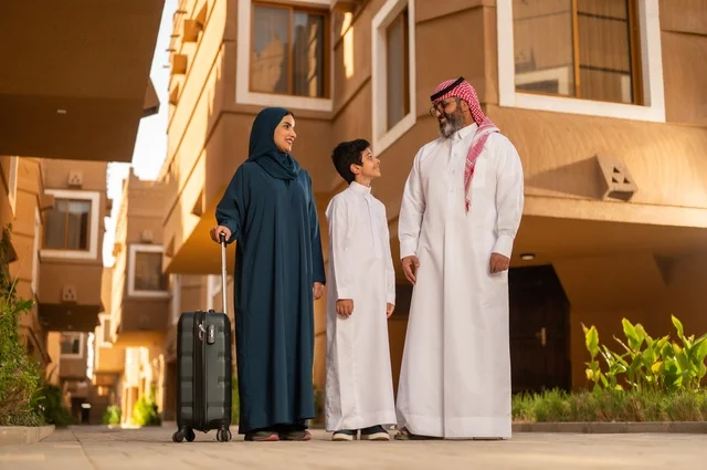 Spending an entertaining summer trip, the atmosphere of the Gulf family and family bonding, an Arab Gulf Saudi family consisting of a father, a mother, and a child standing at the entrance of a modern house. The father is wearing a white thobe with a ghutrah and agal, while the child is wearing a traditional white thobe. The mother appears in an abaya and hijab, holding a travel bag in her hand.
