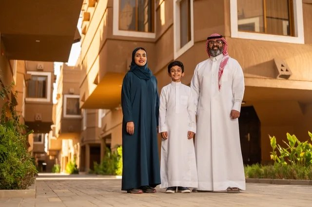 قضاء الأوقات السعيدة مع الابناء، جو العائلة الخليجية والترابط الأسري، عائلة عربية خليجية سعودية مكونة من أب وأم وطفل يقفون في مدخل منزل حديث، يرتدي الأب الثوب الأبيض مع الغترة والعقال، بينما يرتدي الطفل ثوبًا أبيض تقليديًا، وتظهر الأم بعباءة وحجاب