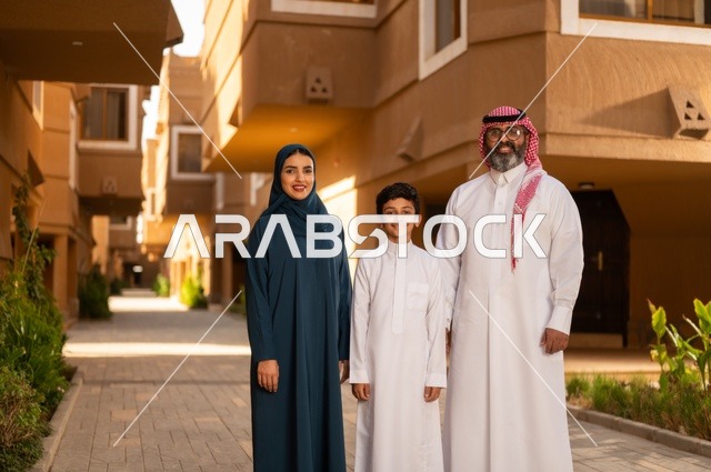 The atmosphere of the Gulf family and family bonding, an Arab Gulf Saudi family consisting of a father, mother, and child standing at the entrance of a modern home. The father is wearing a white thobe with a ghutrah and agal, while the child is dressed in a traditional white thobe, and the mother appears in a black abaya and hijab.